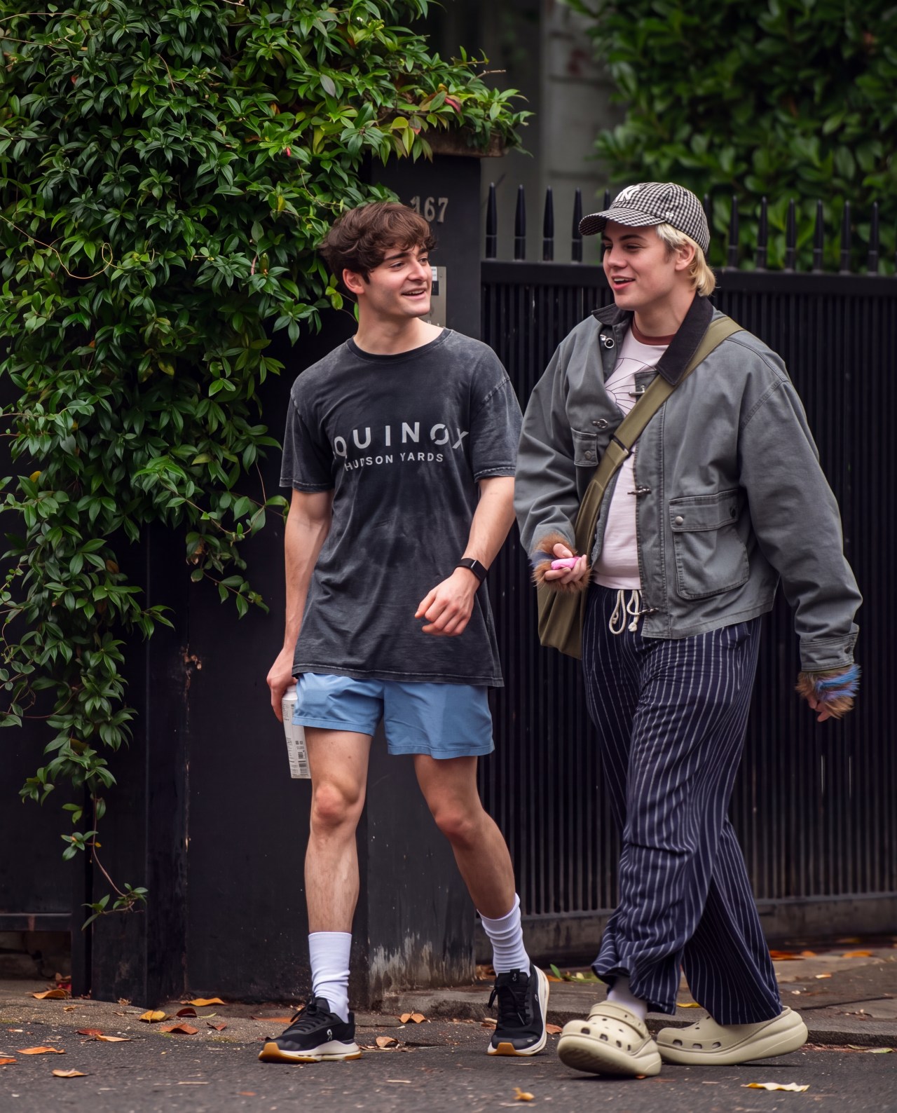 Two young men walking and smiling on a city sidewalk; one in a dark Equinox tee and blue shorts, the other in a grey jacket, pinstripe trousers, and Crocs.