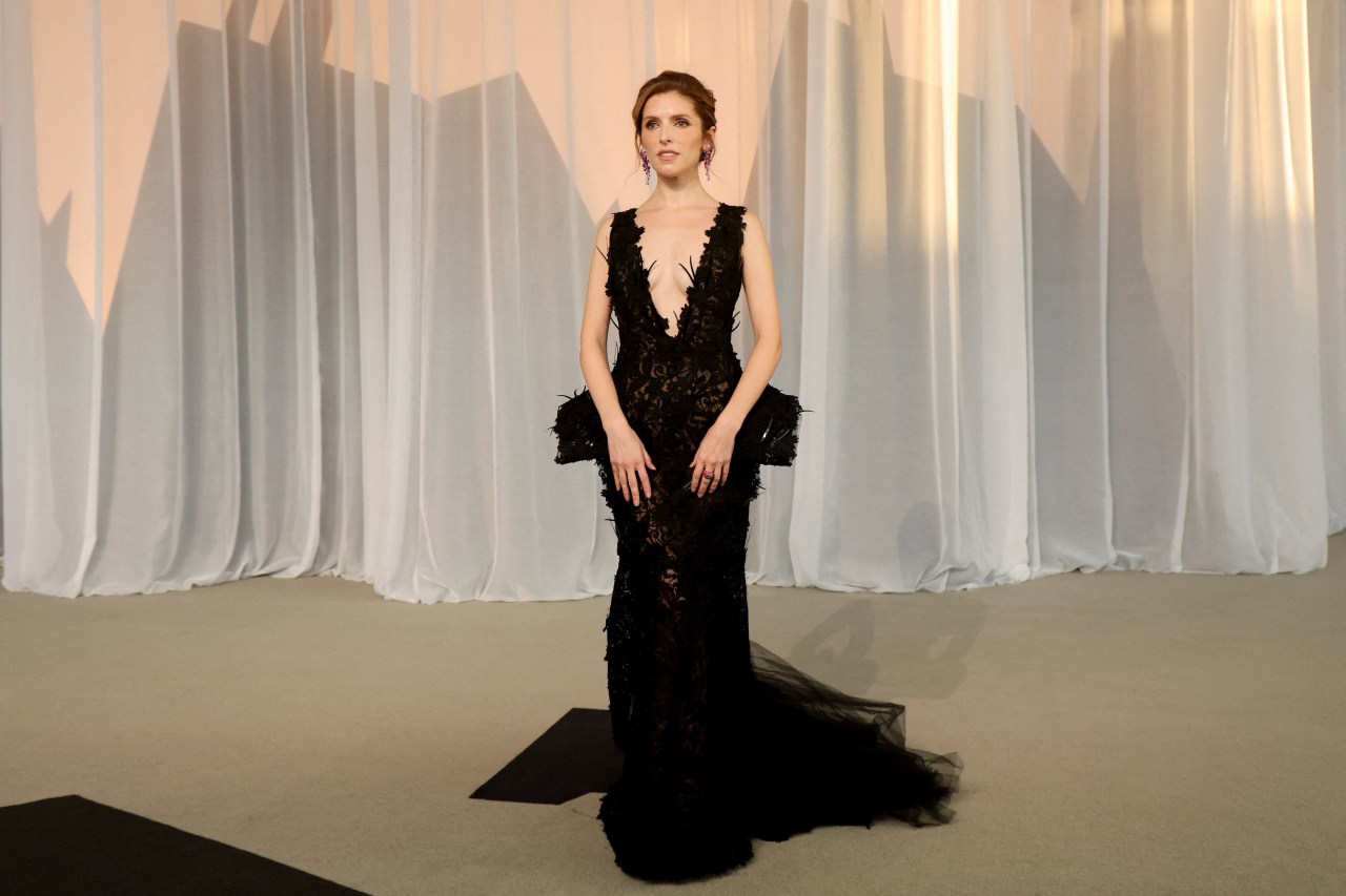 Woman in a plunging black lace gown with a feathered peplum and long train, standing in front of draped white curtains at a formal event.