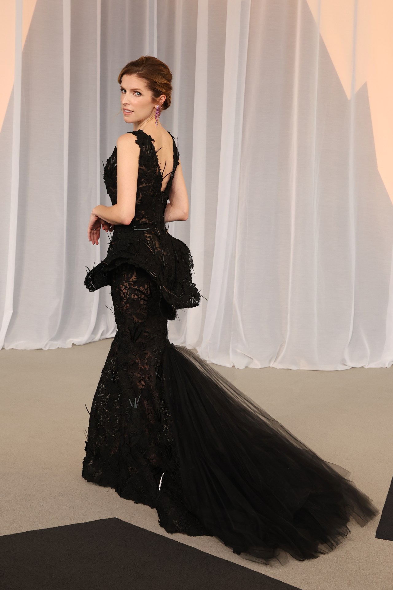 Woman in a black lace mermaid gown with feather details and a tulle train, photographed from behind as she looks back over her shoulder.