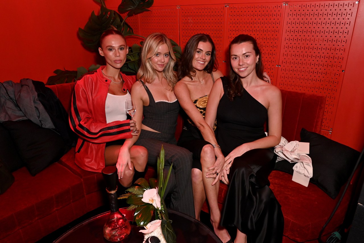 Four women seated on a red sofa in a red-lit venue; the woman on the far left wears a red leather jacket and holds a wine glass.
