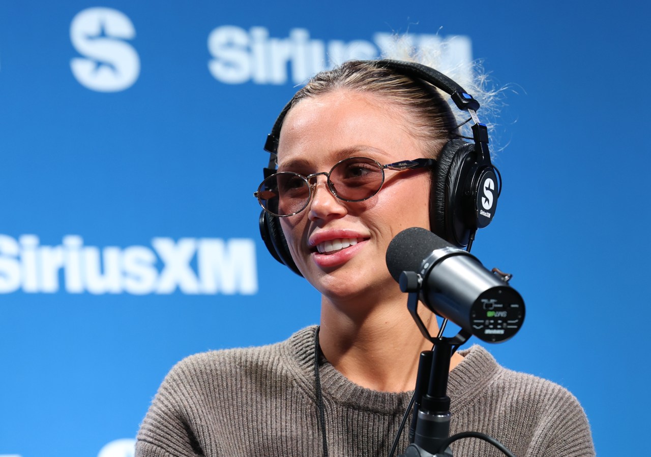Close-up of a smiling woman wearing tinted Prada glasses and SiriusXM headphones in front of a blue SiriusXM backdrop, leaning toward a studio microphone.