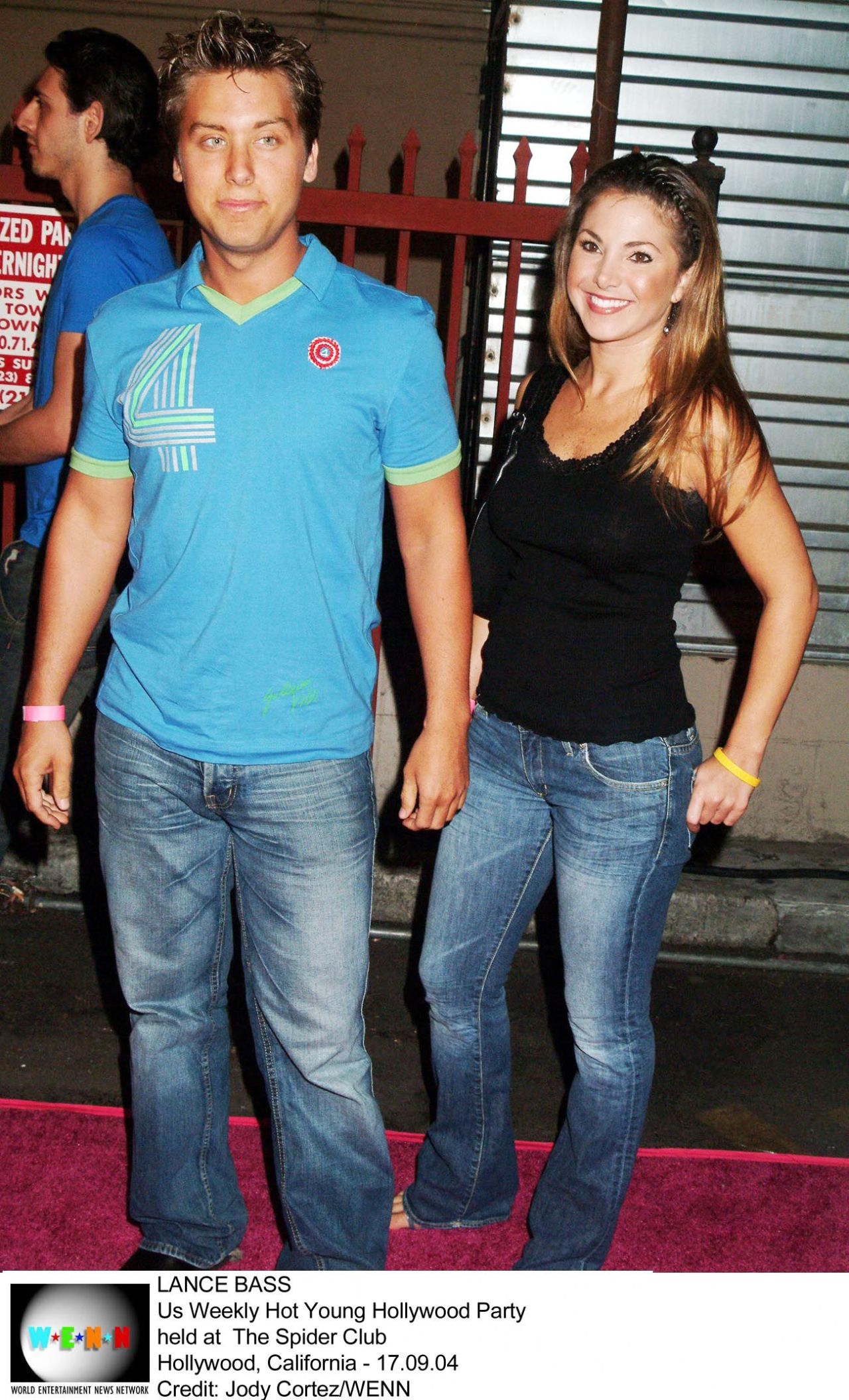 Lance Bass in a blue polo shirt and jeans standing next to a brunette woman in a black top and jeans on a pink carpet outside a venue.