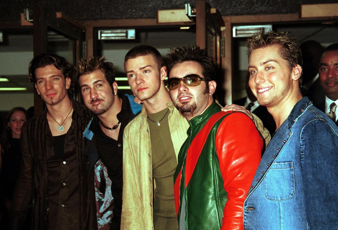 Five members of NSYNC standing together indoors at an event, wearing early-2000s fashion including a green and red leather jacket and a denim jacket.
