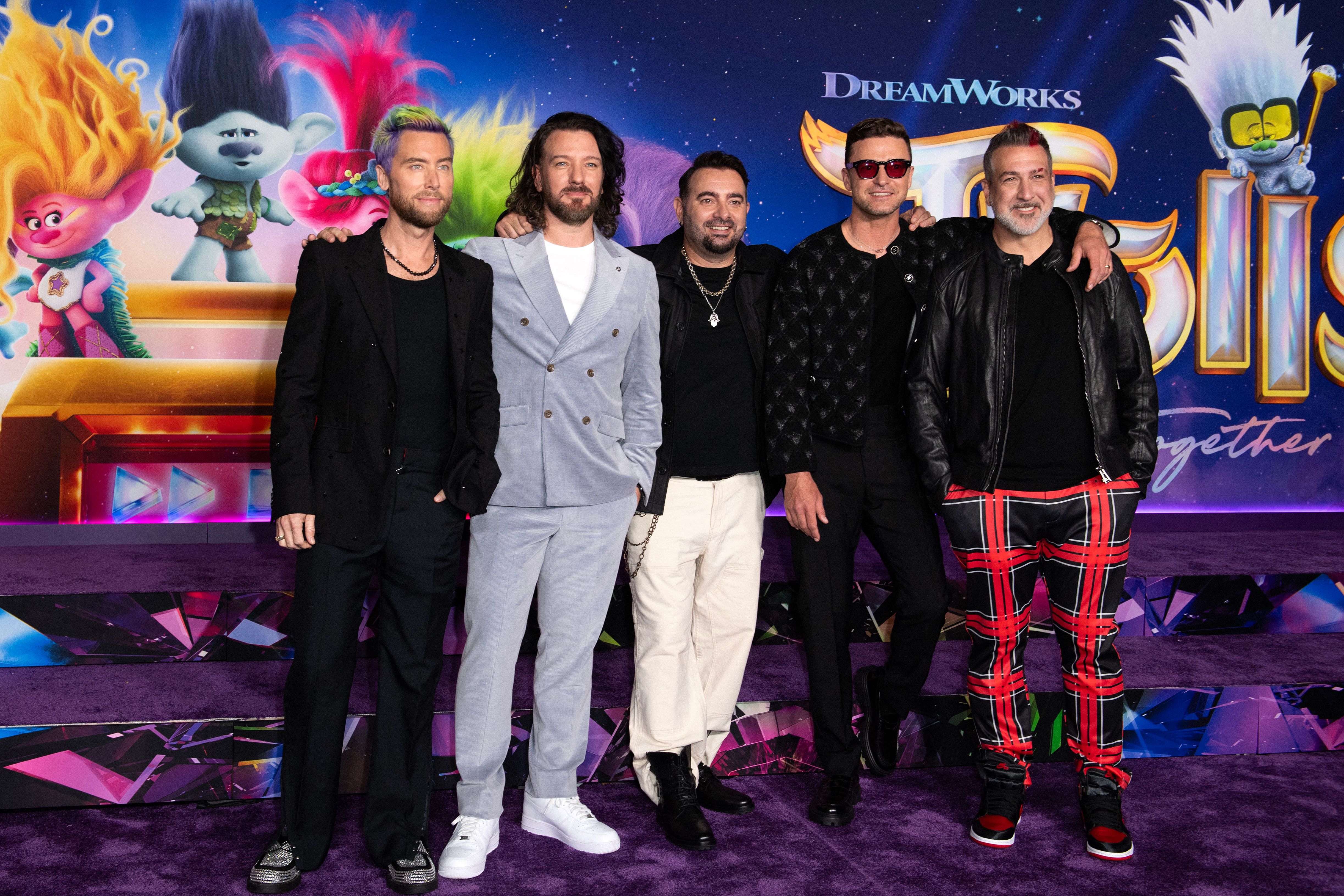 Five men standing together in front of a DreamWorks Trolls: Band Together premiere backdrop, dressed in a mix of black outfits and one grey suit.