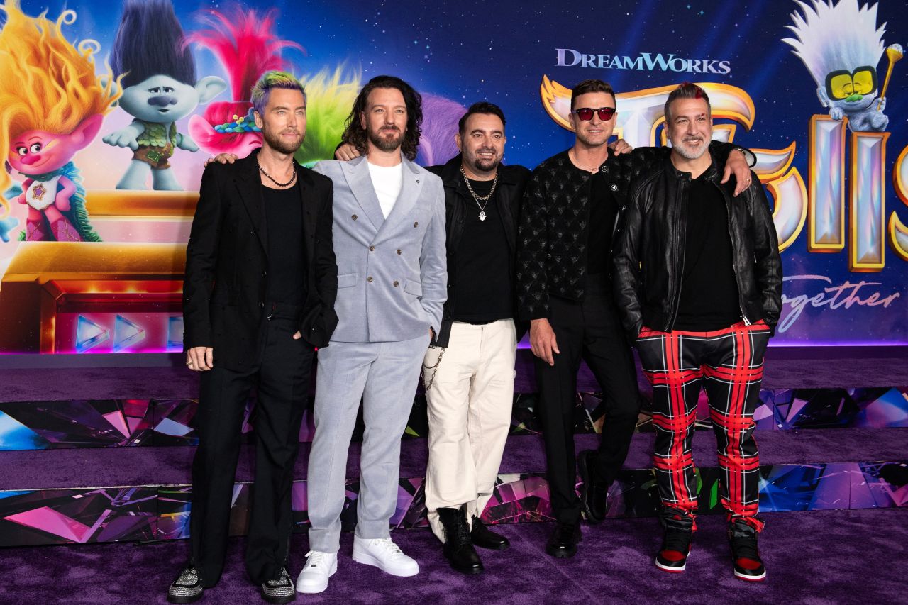 Five men standing together in front of a DreamWorks Trolls: Band Together premiere backdrop, dressed in a mix of black outfits and one grey suit.