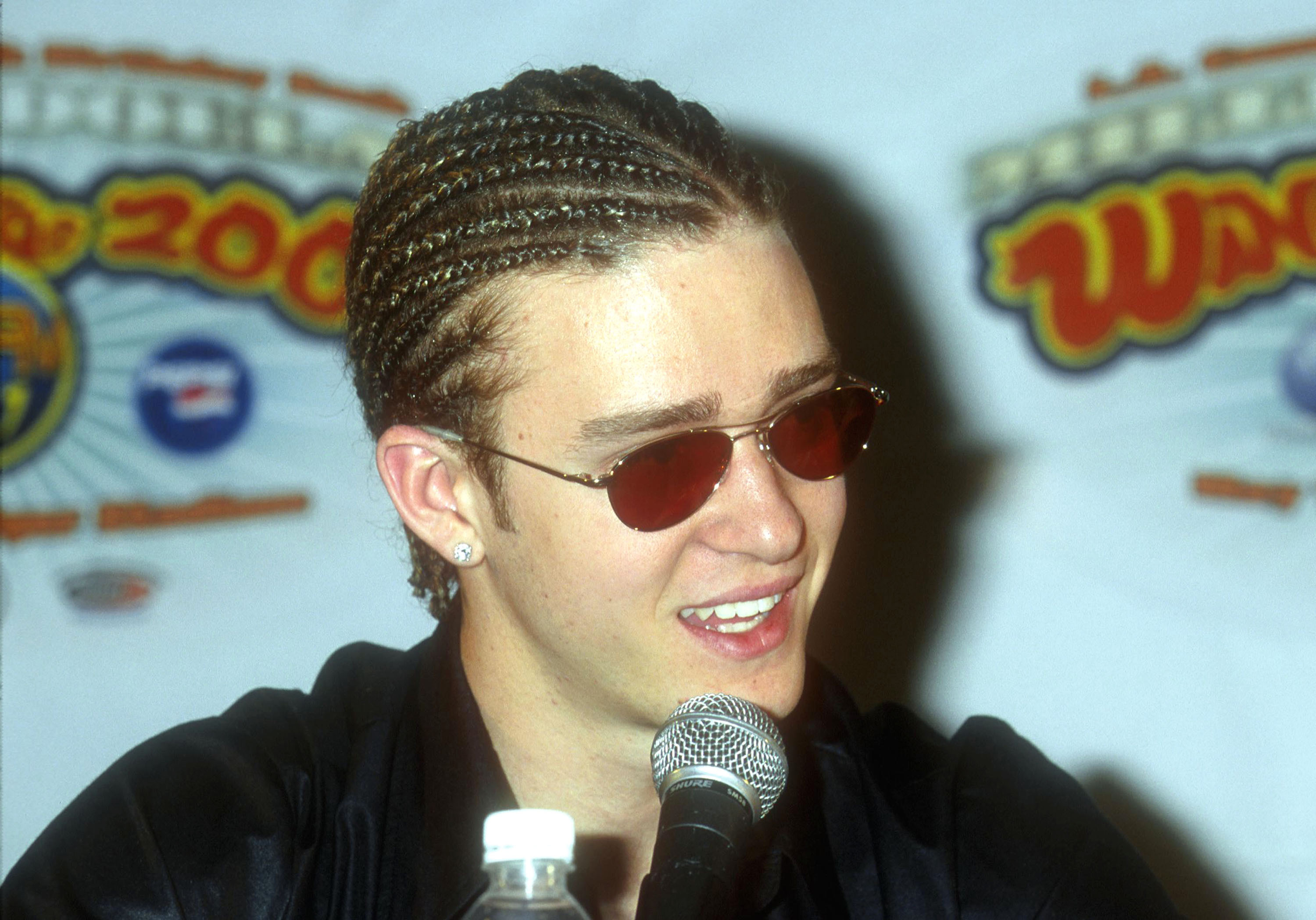 Young man with cornrows and red-tinted aviator sunglasses speaking into a microphone at a press event.