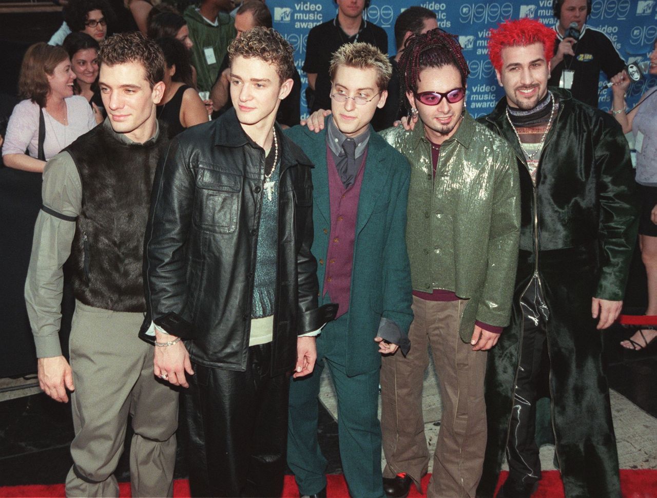 Five members of NSYNC pose together on the red carpet at the 1999 MTV Video Music Awards, dressed in late-90s fashion including leather jackets and a green metallic coat.