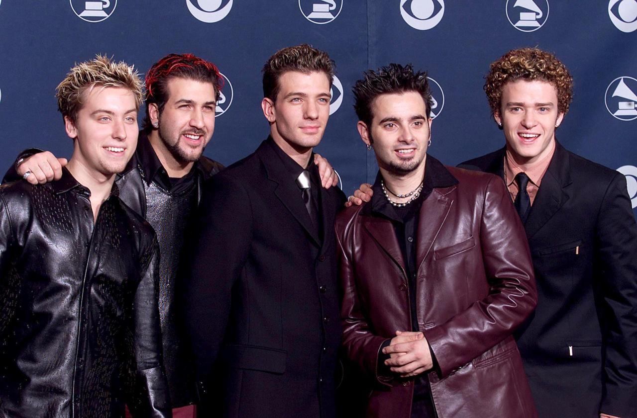 Five young men in stylish jackets and suits posing together at the Grammy Awards, smiling at cameras against a Grammy logo backdrop.