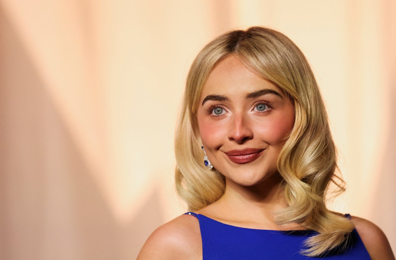Close-up of Sabrina Carpenter smiling in a cobalt blue gown and sapphire drop earrings against a warm peach backdrop.