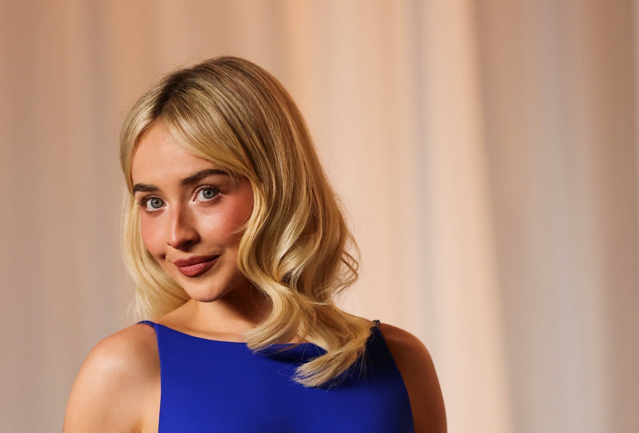 Sabrina Carpenter in a royal blue sleeveless gown, looking over her shoulder with a slight smile, draped curtains in background.