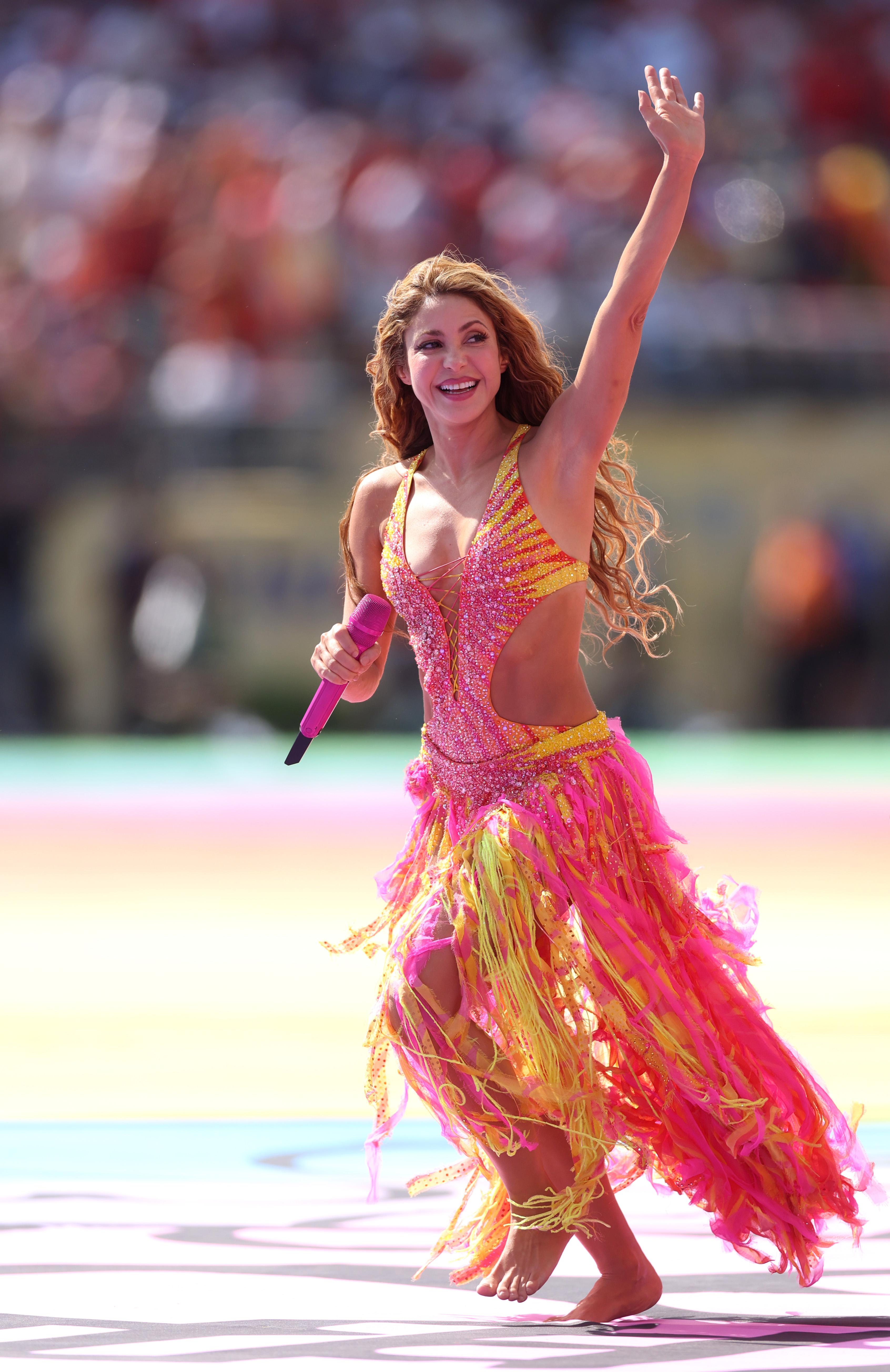 Shakira in a pink and yellow beaded fringe costume, holding a microphone and raising one arm while performing barefoot at a stadium event.