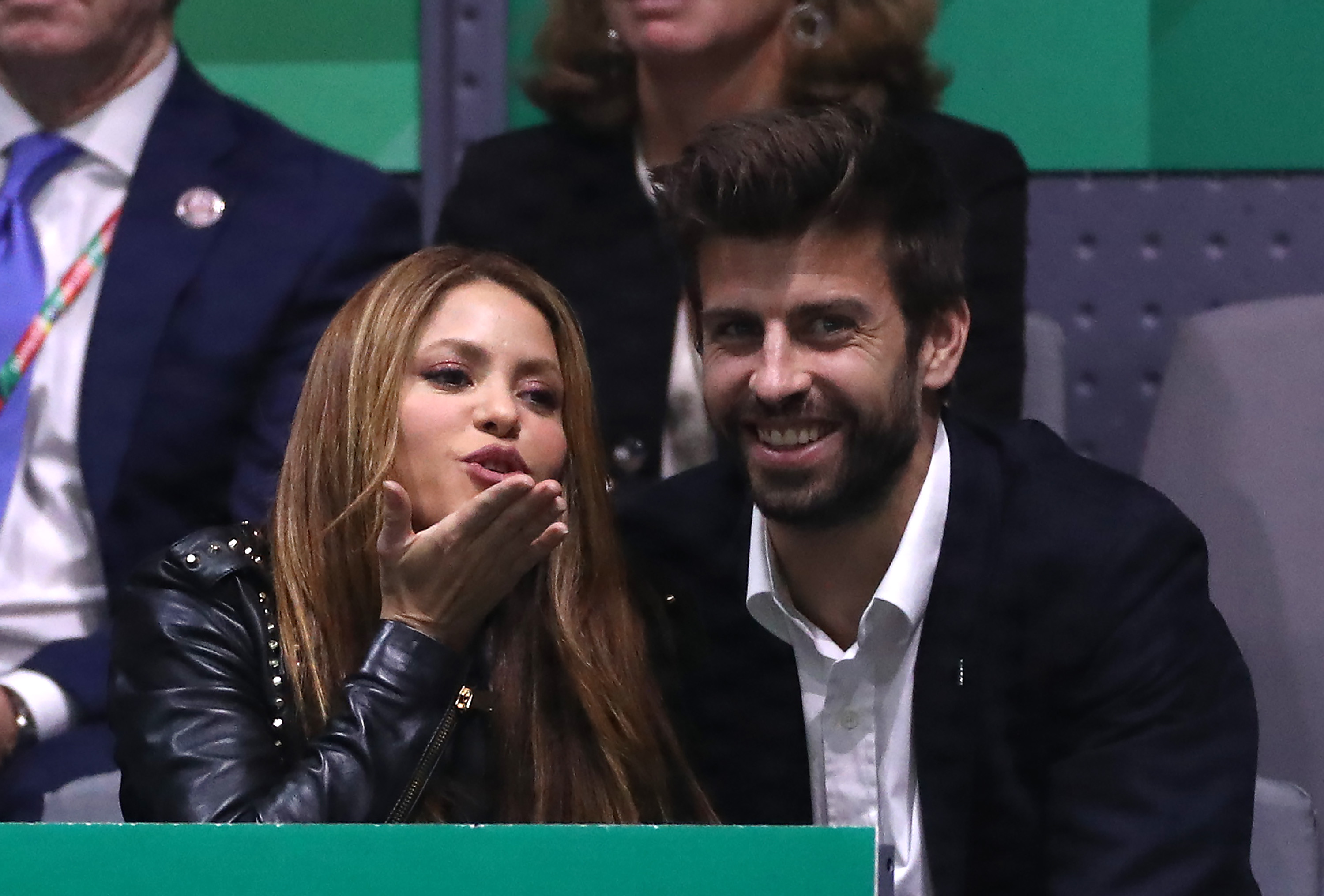 Shakira blowing a kiss while Gerard Piqué smiles beside her in stadium seating at a tennis event.